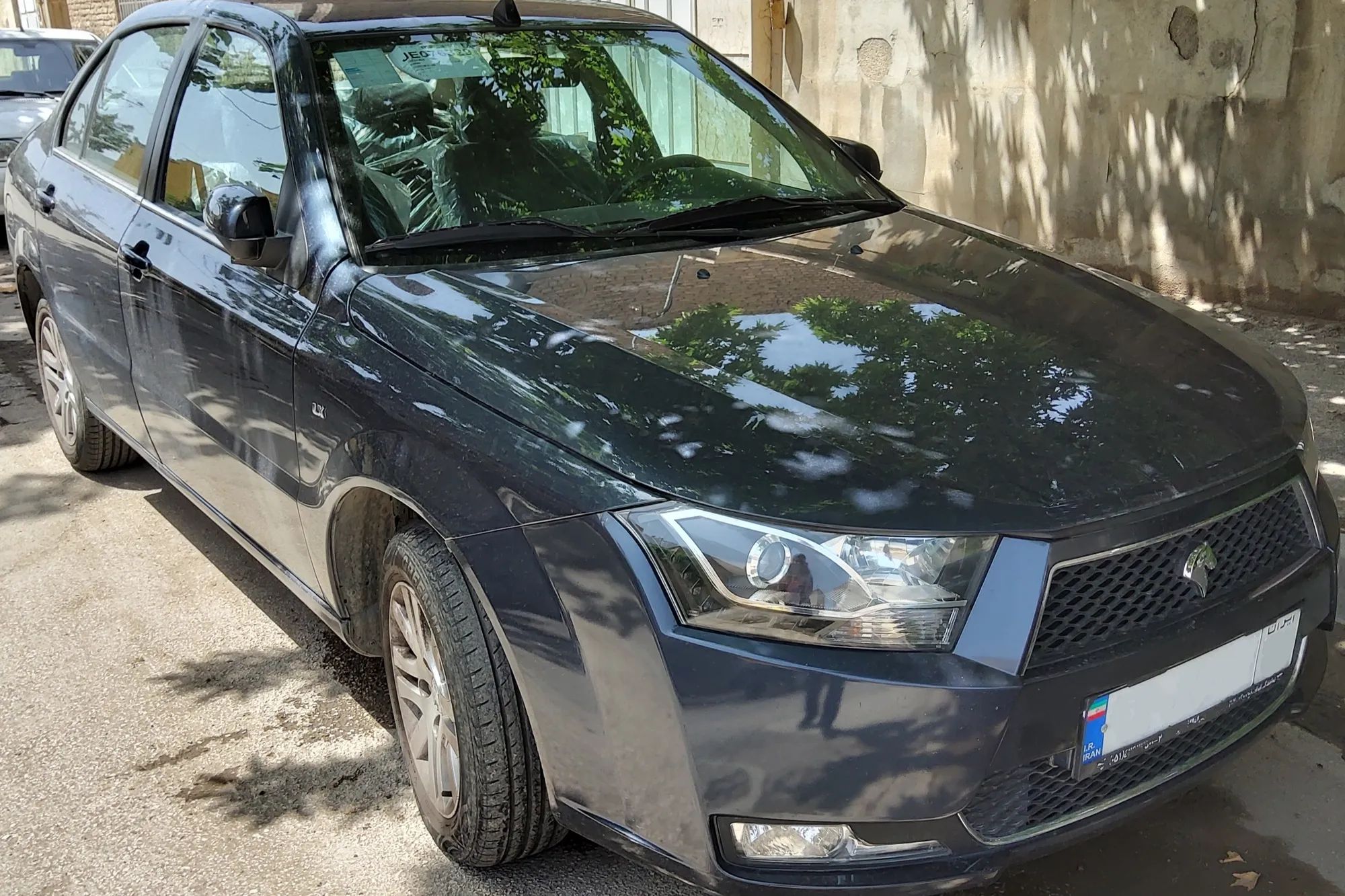 دنا ال‌ایکس آبی تیره با جلوپنجره سیاه، نمای جلو و کنار، پارک‌شده کنار دیواری در سایه درختان